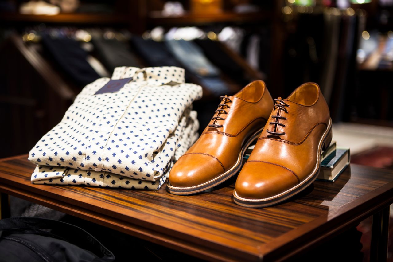 services-05 Elegant brown leather shoes and printed shirt displayed on a wooden table in a stylish clothing store.