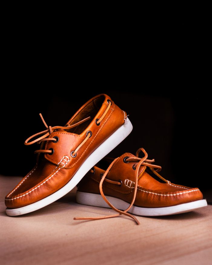services-06 Close-up of brown leather boat shoes with white soles on a wooden floor against a black background.